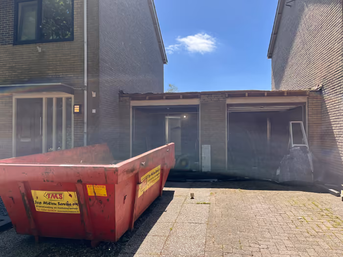 Twee bestaande garages met gestripte gevelopeningen en een rode afvalcontainer ervoor