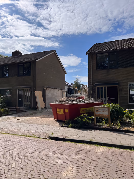 Volle puincontainer en bouwbord op straat voor twee woningen tijdens de sloop