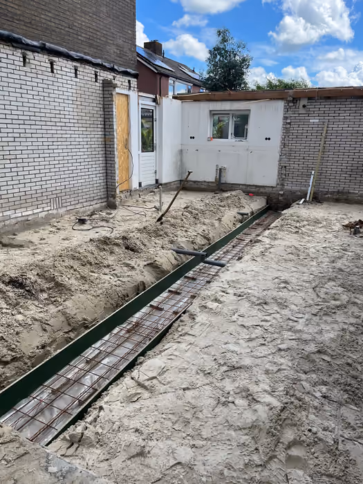 Uitgegraven funderingssleuf met wapeningskorf en rioleringsbuizen in het zand