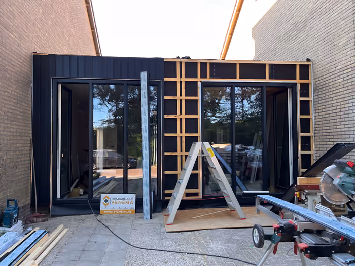 Aanbouw met zwarte kozijnen, gevelbekleding deels geplaatst op houten rachelwerk