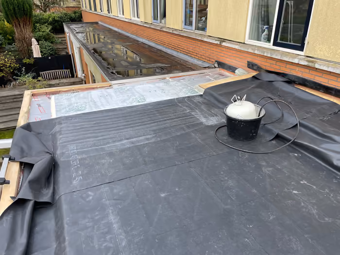 Zwarte dakbedekking uitgerold op een plat dak naast een glazen overkapping