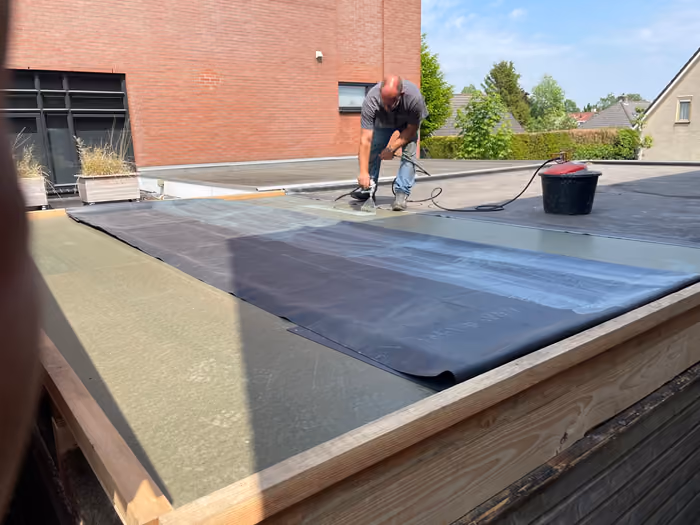 Man rolt zwarte dakbedekking uit over isolatieplaten op een plat dak