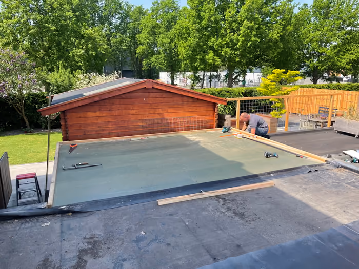 Timmerman op een plat dak legt isolatieplaten naast een houten tuinhuis