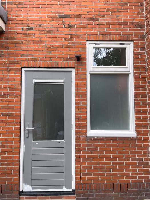 Grijze achterdeur met glasvlak en wit draaikiepraam in een bakstenen gevel