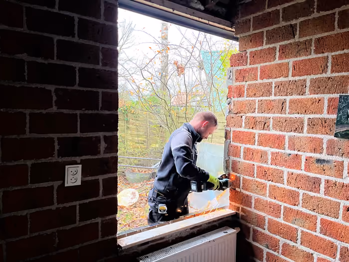 Timmerman zaagt in een gevelopening, gezien vanuit de kale kamer binnen