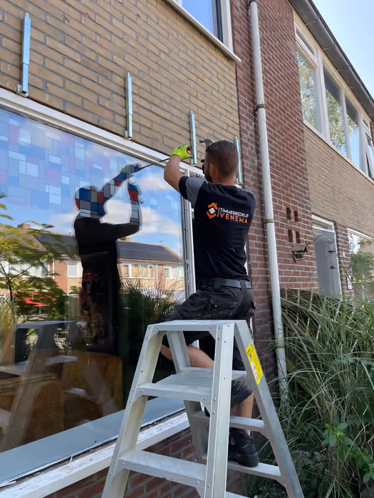 Timmerman met bedrijfsshirt monteert een groot raam vanaf een trapladder