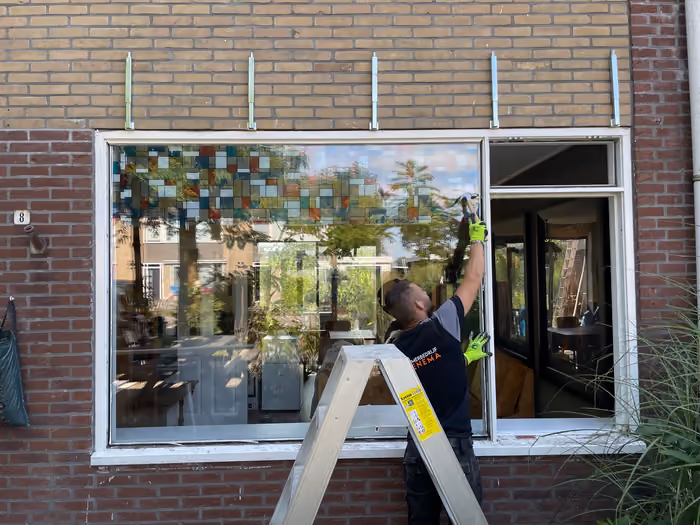 Timmerman op een trapladder werkt aan een groot raamkozijn in de gevel