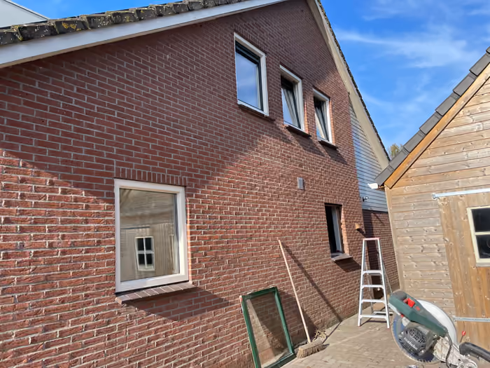 Zijgevel met nieuwe kozijnen bij blauwe lucht, ladder en oud kozijn ernaast