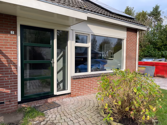Voorgevel met nieuw raamkozijn en donkergroene deur, afvalcontainer naast het huis