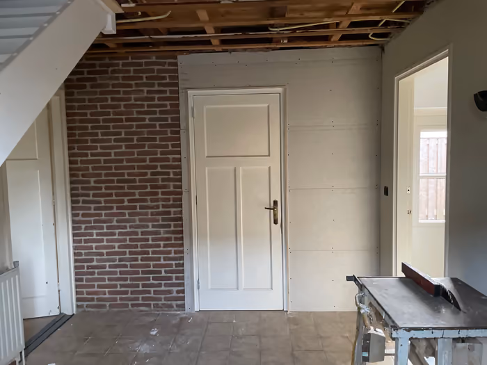 Hal met bakstenen wand, witte binnendeur en plafond gestript tot de balken