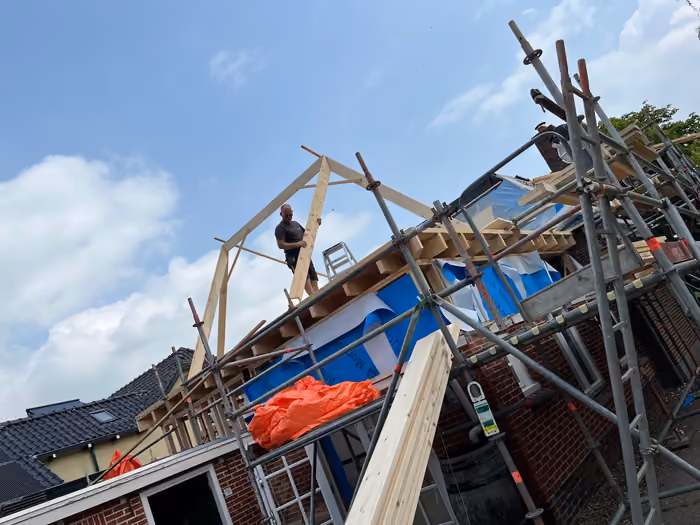 Timmerman tilt een houten spant omhoog op de steiger tegen blauwe lucht