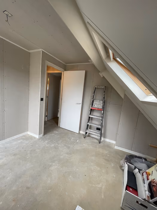 Zolderkamer met dakraam, witte deur en trapje tegen de gipsplaten wand