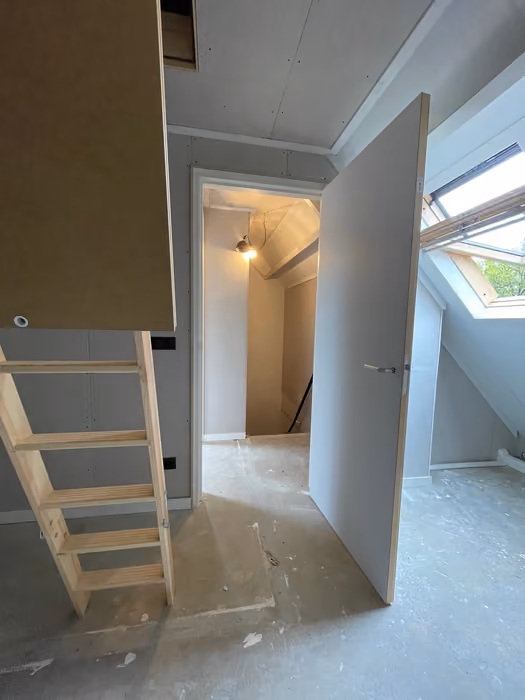 Houten trap naar de zolder, openstaande deur en dakraam met groen uitzicht